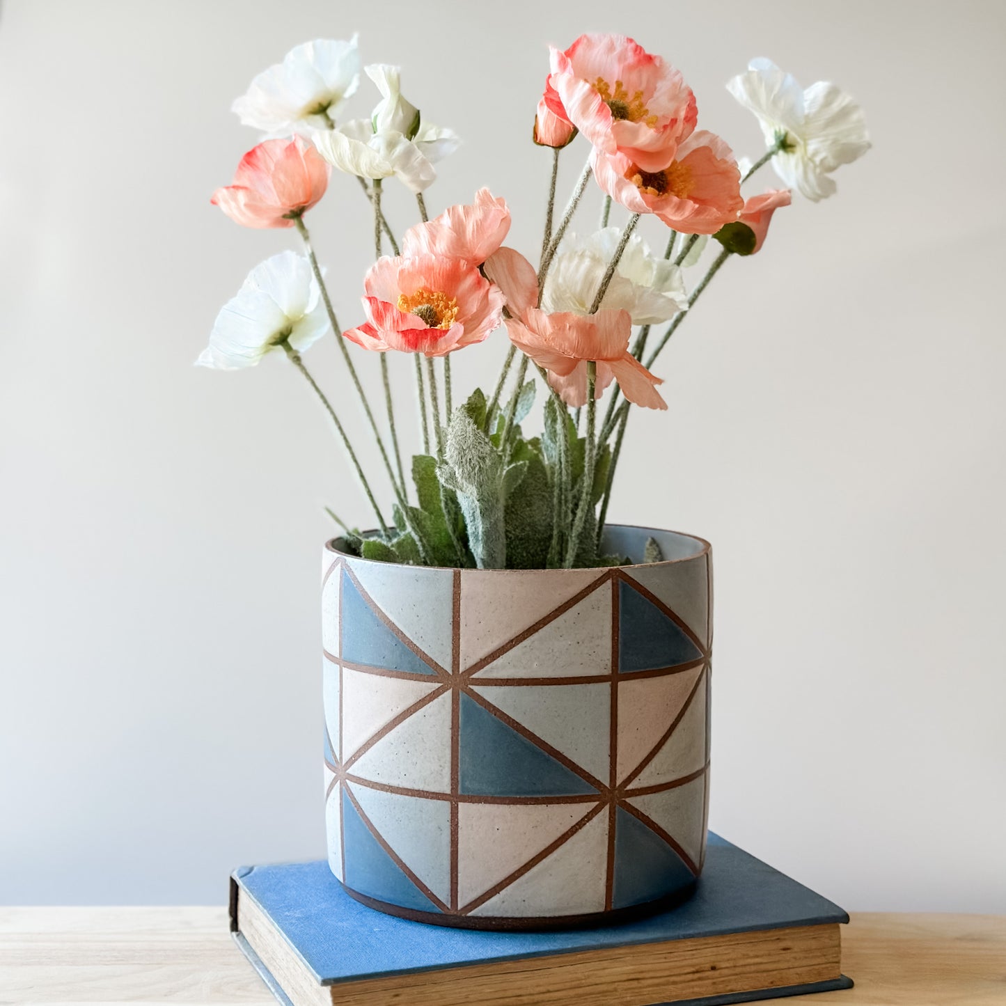 A geometric handmade ceramic planter pot sits on a book and wooden table. The planter pot has flowers and is painted with dark blue, pink, grey and white glaze.