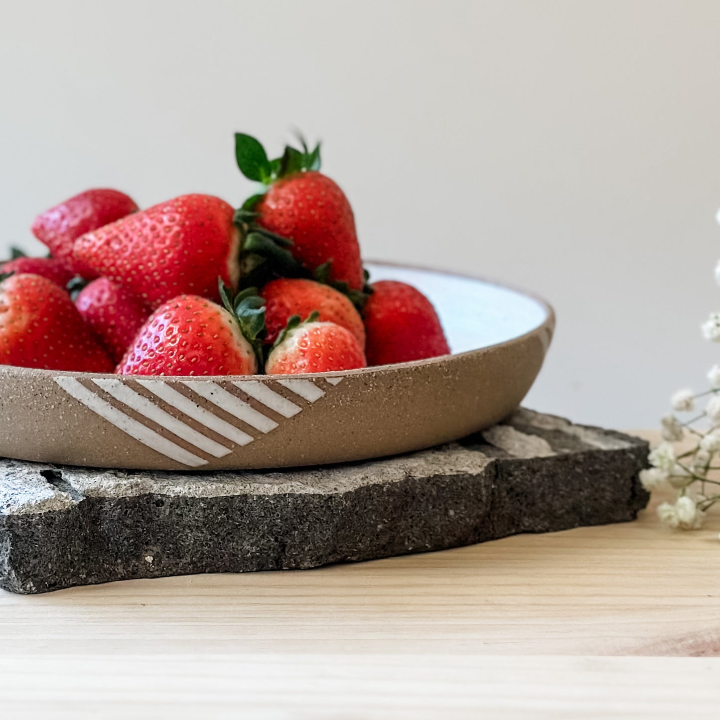 A close-up view of a Geometric Handmade Ceramic Platter with strawberries. The interior of the platter has a white glaze and the exterior has natural clay exposed to show a geometric pattern. The handmade platter sits on a tile and a wooden table.