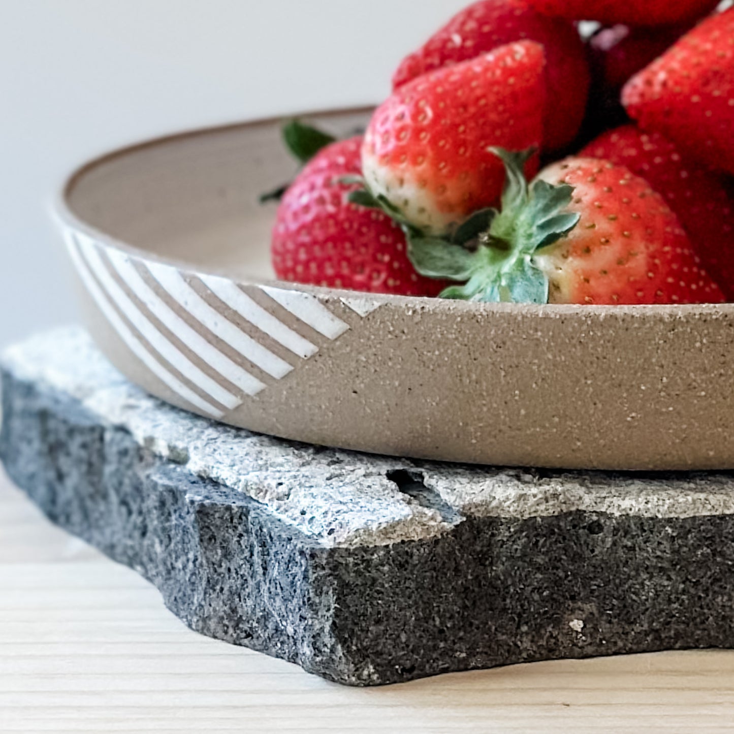 A close-up view of a Geometric Handmade Ceramic Platter with strawberries. The interior of the platter has a white glaze and the exterior has natural clay exposed to show a geometric pattern. The handmade platter sits on a tile and a wooden table.