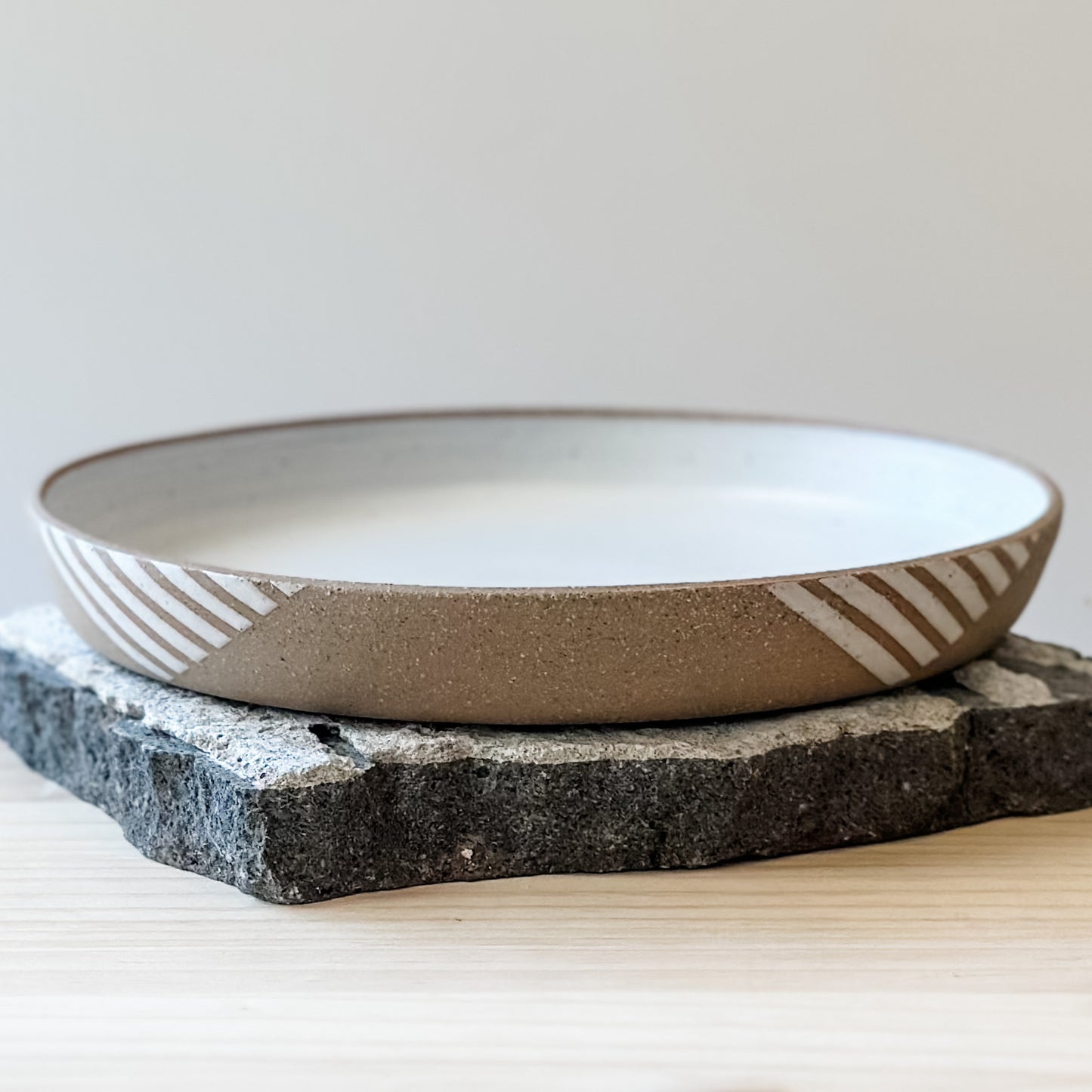 A front view of a Geometric Handmade Ceramic Platter in a white glaze. The exterior of the platter has natural clay exposed to show a geometric pattern. The handmade platter sits on a tile and a wooden table.