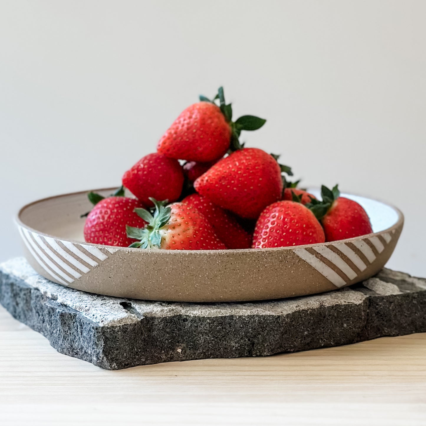 A front view of a Geometric Handmade Ceramic Platter with strawberries. The interior of the platter has a white glaze and the exterior has natural clay exposed to show a geometric pattern. The handmade platter sits on a tile and a wooden table.