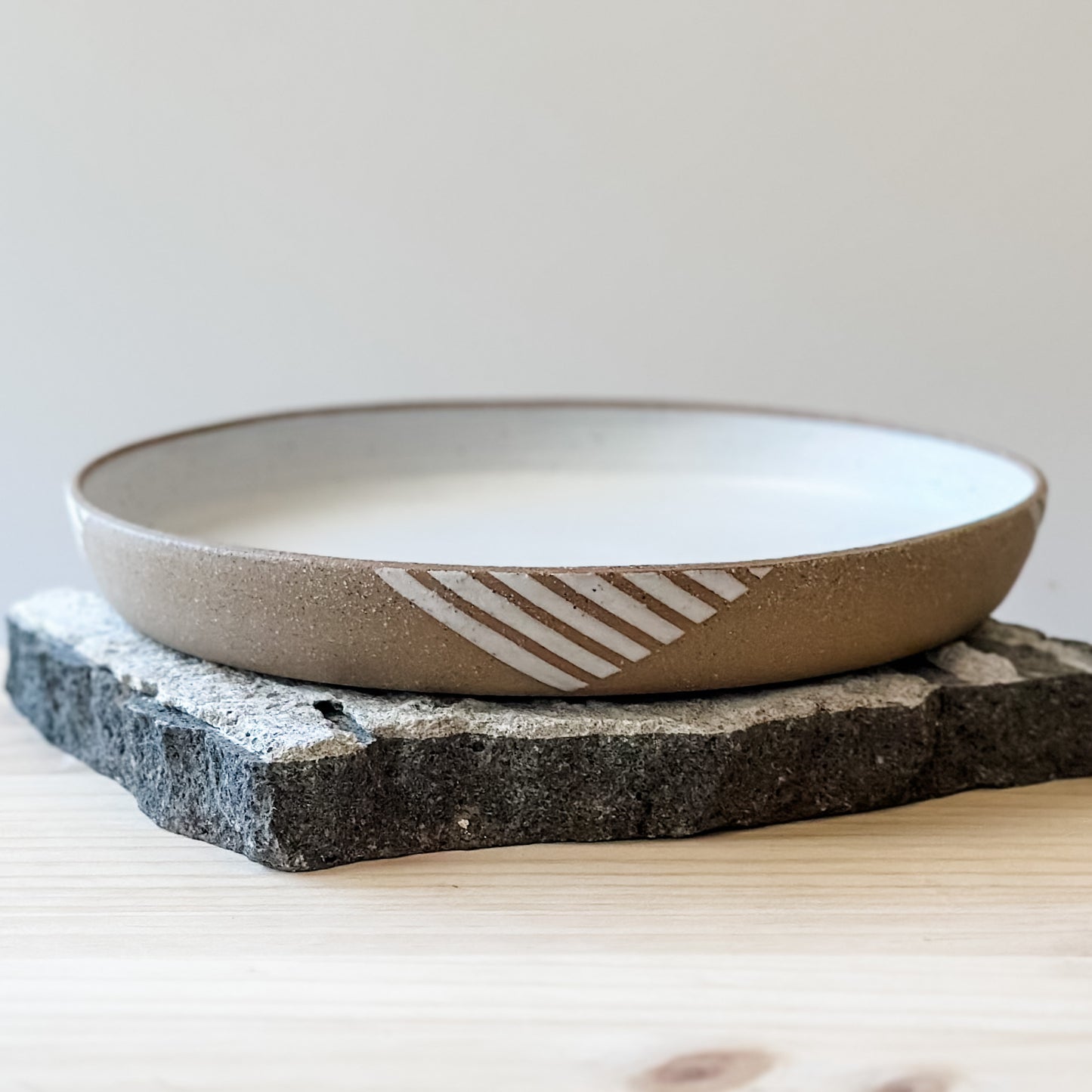 A front view of a Geometric Handmade Ceramic Platter in a white glaze. The exterior of the platter has natural clay exposed to show a geometric pattern. The handmade platter sits on a tile and a wooden table.
