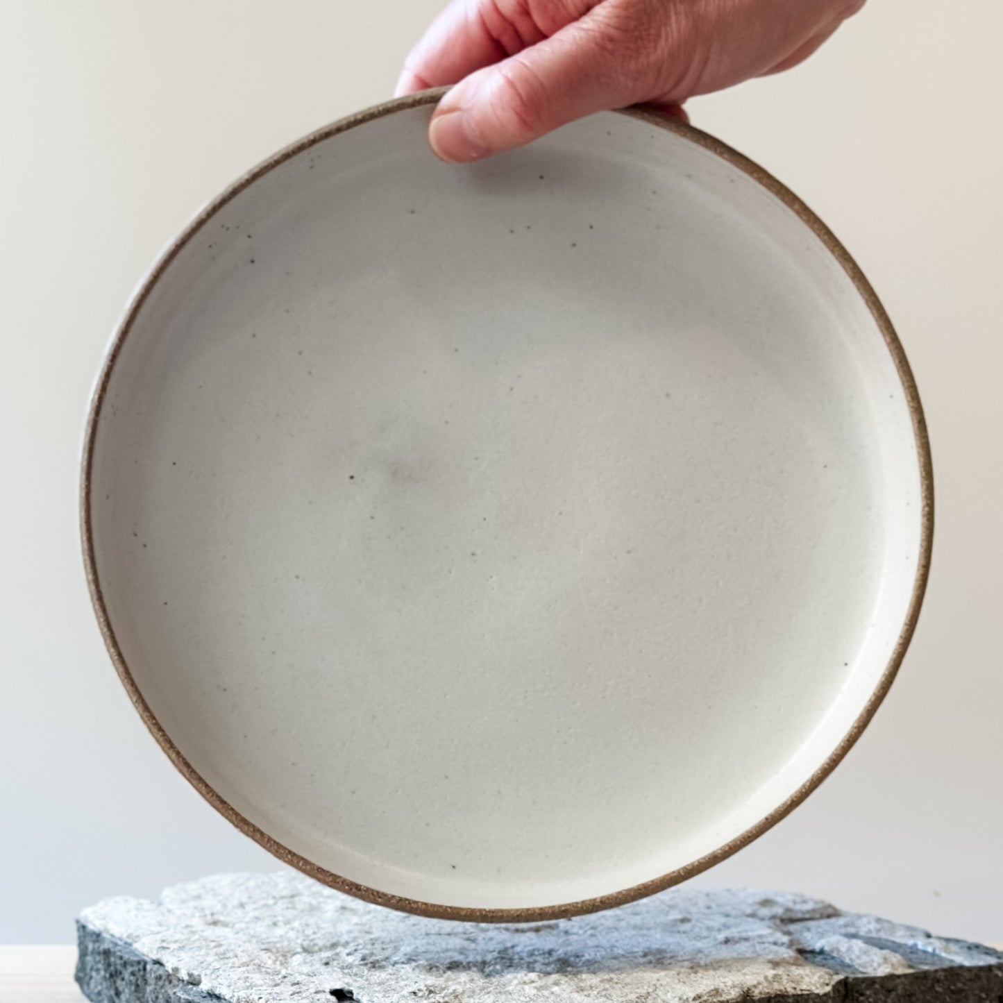 A interior view of the Geometric Handmade Ceramic Platter. The interior of the platter has white glaze with natural clay on the rim. The handmade platter is being held with one hand above a tile.