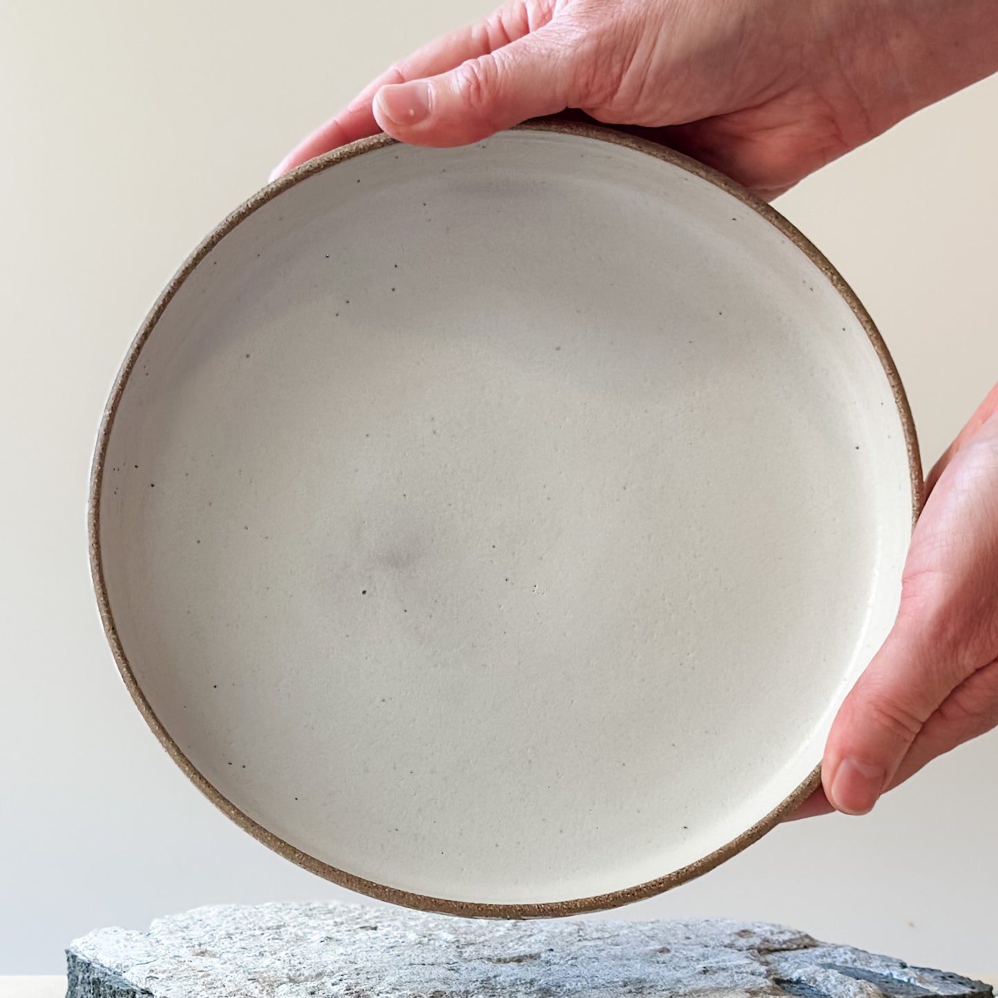 A interior view of the Geometric Handmade Ceramic Platter. The interior of the platter has white glaze with natural clay on the rim. The handmade platter is being held with two hands above a tile.