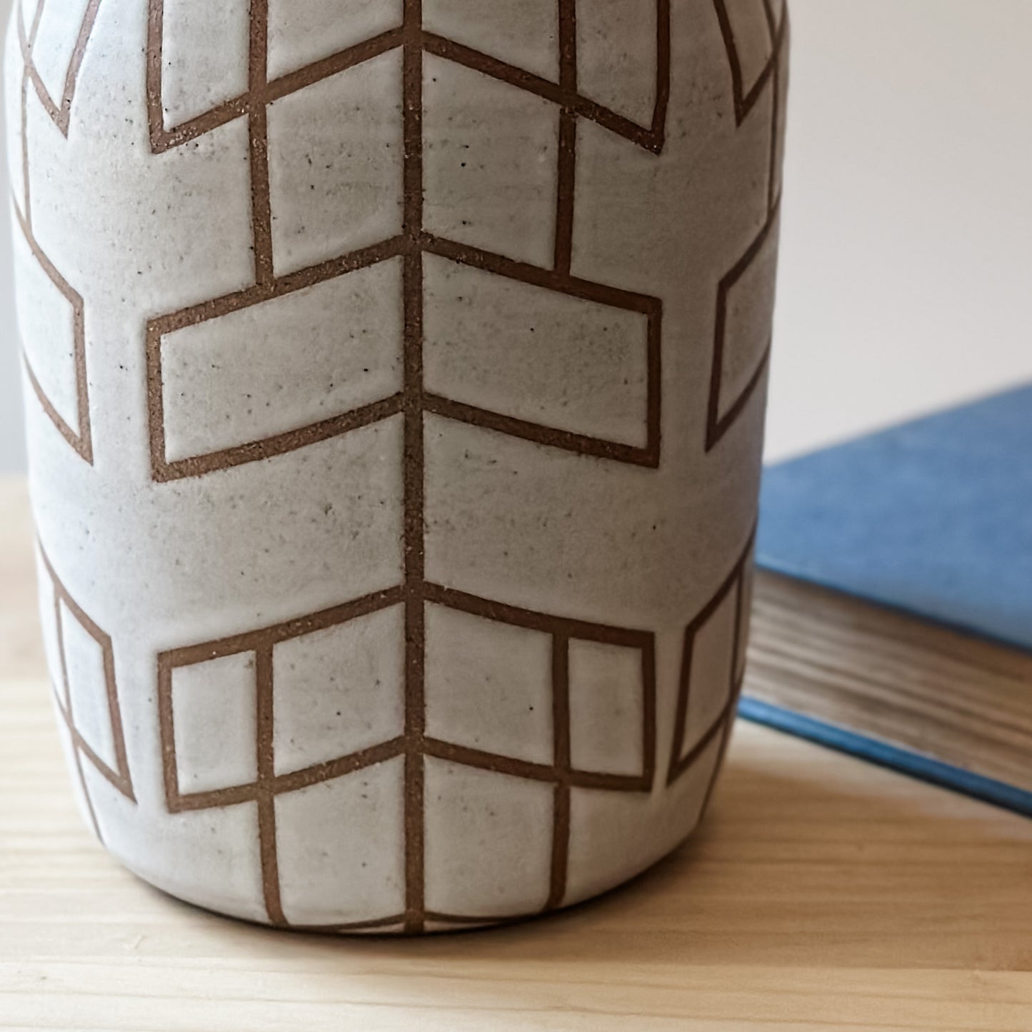 A close up view of a Handmade Geometric Vase. The Vase is glazed white, and the unglazed clay makes the pattern. The handmade vase sits next to a book on a wooden table.