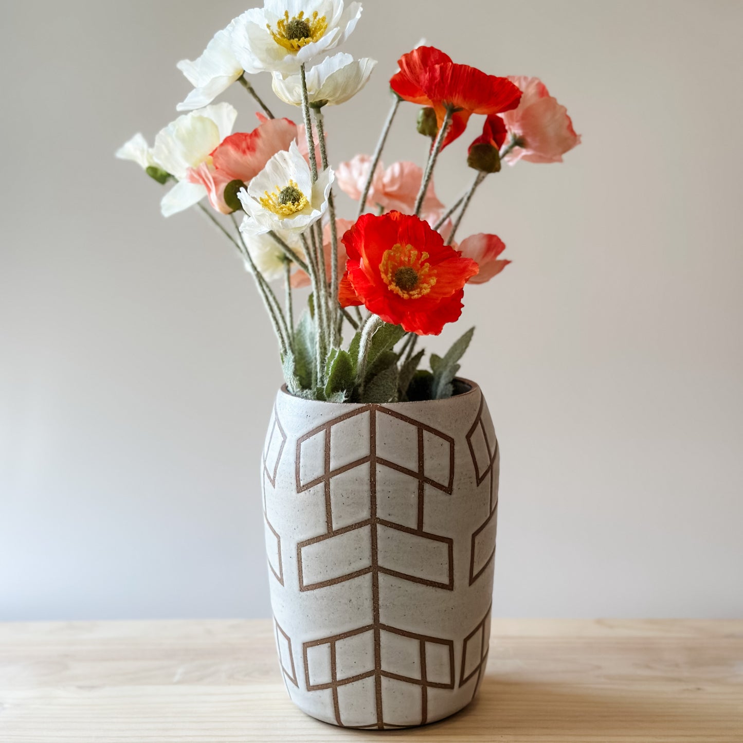 A Geometric Handmade Ceramic vase sits on a wooden table. The Geometric Ceramic Vase is glazed white. The pattern is made by the unglazed clay. The vase contains a bunch of flowers.