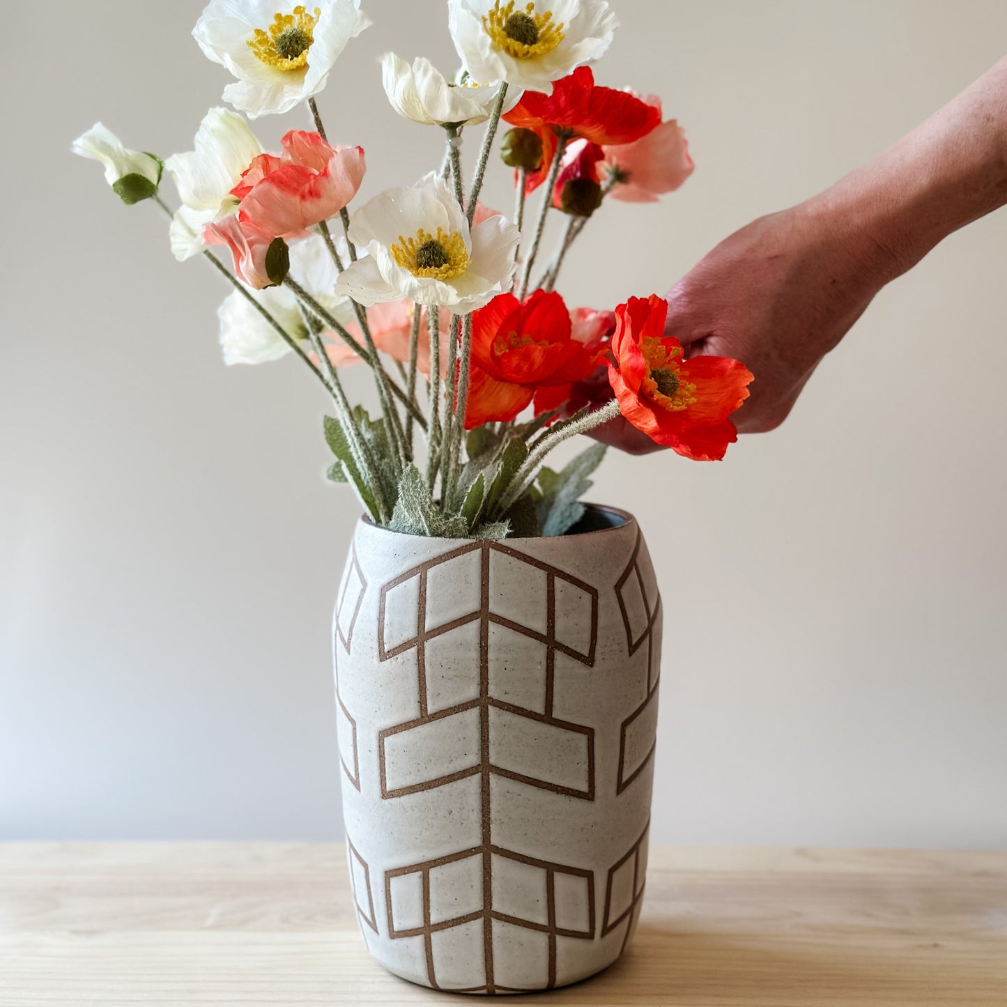 A Geometric Handmade Ceramic vase sits on a wooden table. The Geometric Ceramic Vase is glazed white. The pattern is made by the unglazed clay. A hand is arranging the flowers inside the vase.