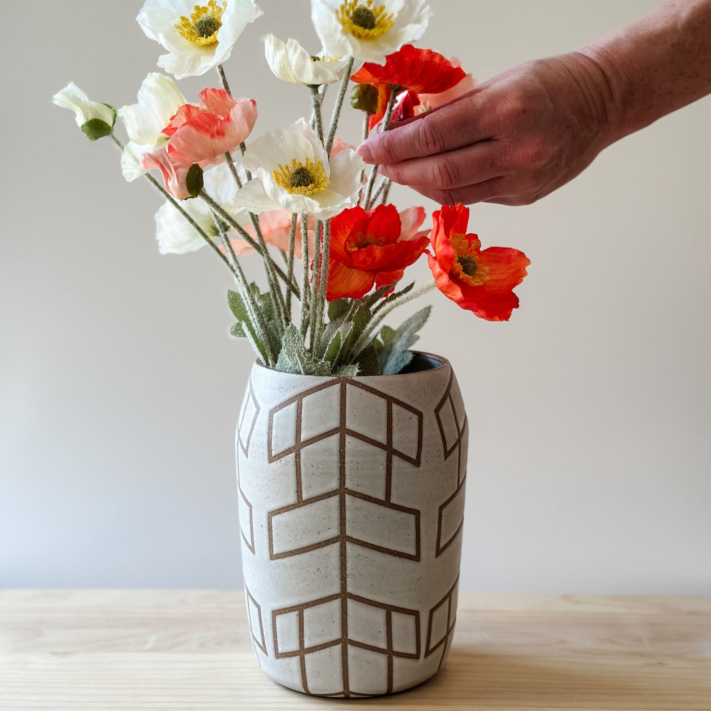 A Geometric Handmade Ceramic vase sits on a table. The Vase is glazed white, and the unglazed clay makes the pattern. A hand arranges the flowers inside the vase.