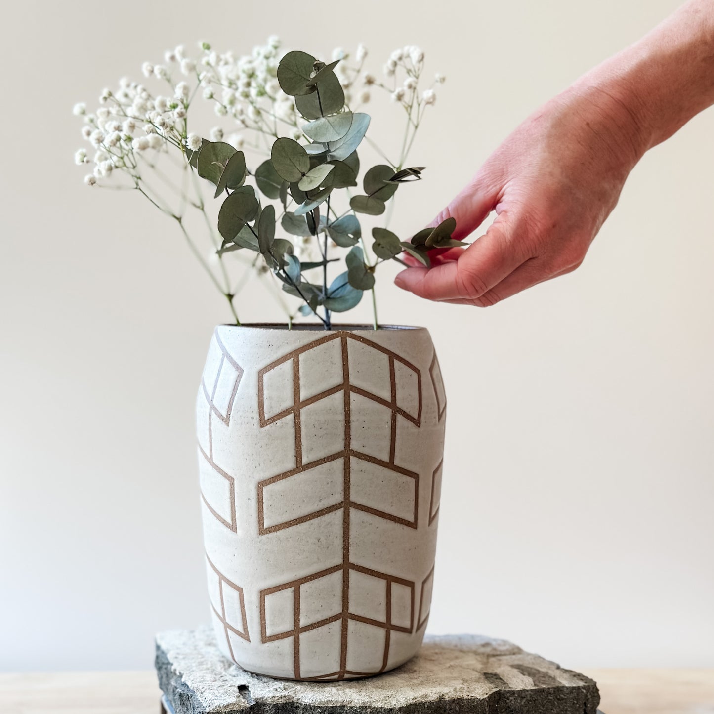 A Geometric Handmade Ceramic vase sits on a tile and on top of a table. The Vase is glazed white, and the unglazed clay makes the pattern. A hand arranges the gum leaves and baby's breath inside the vase.