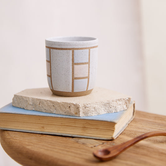A front view of the Geometric Handmade Ceramic Tumbler in soft pink, white and grey glaze. The handmade tumbler sits on a tile, book and wooden table in a coastal-styled setting.