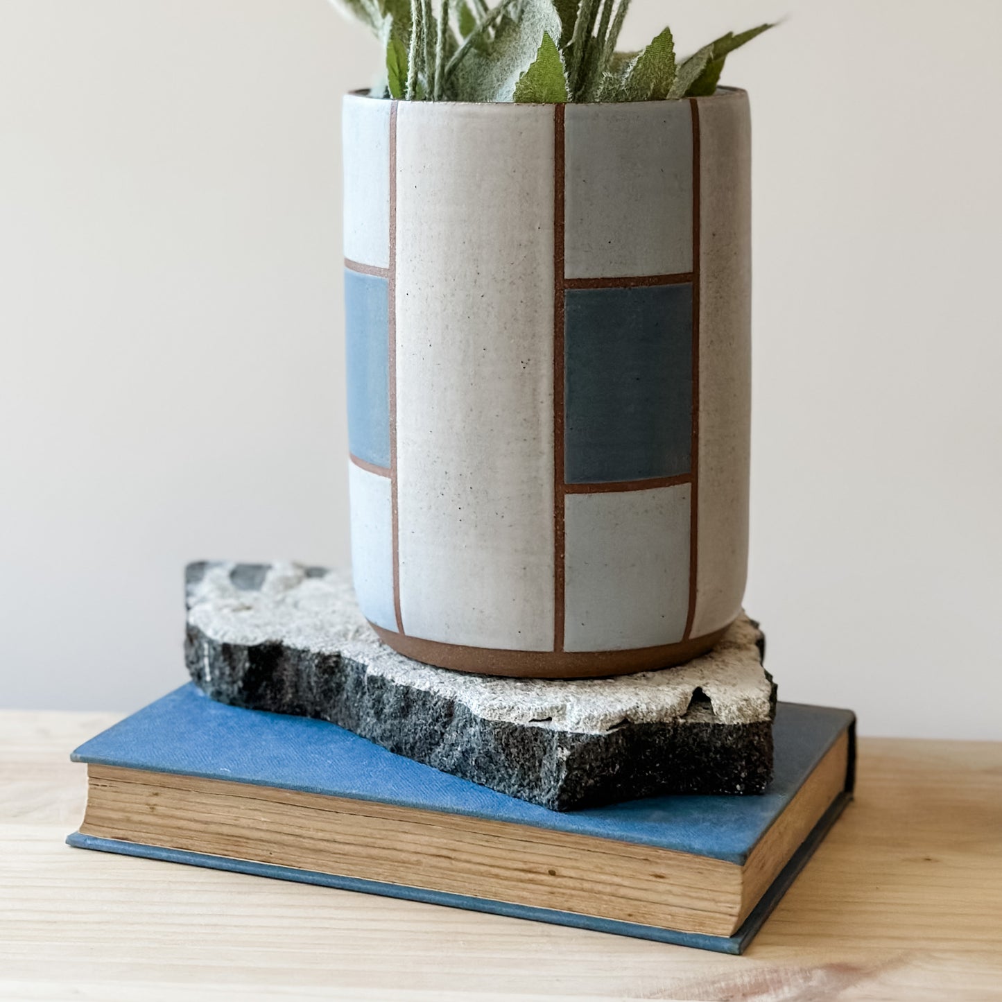 A close up view of a geometric handmade ceramic vase sitting on a tile on a wooden table. The vase is dark blue, grey and white.