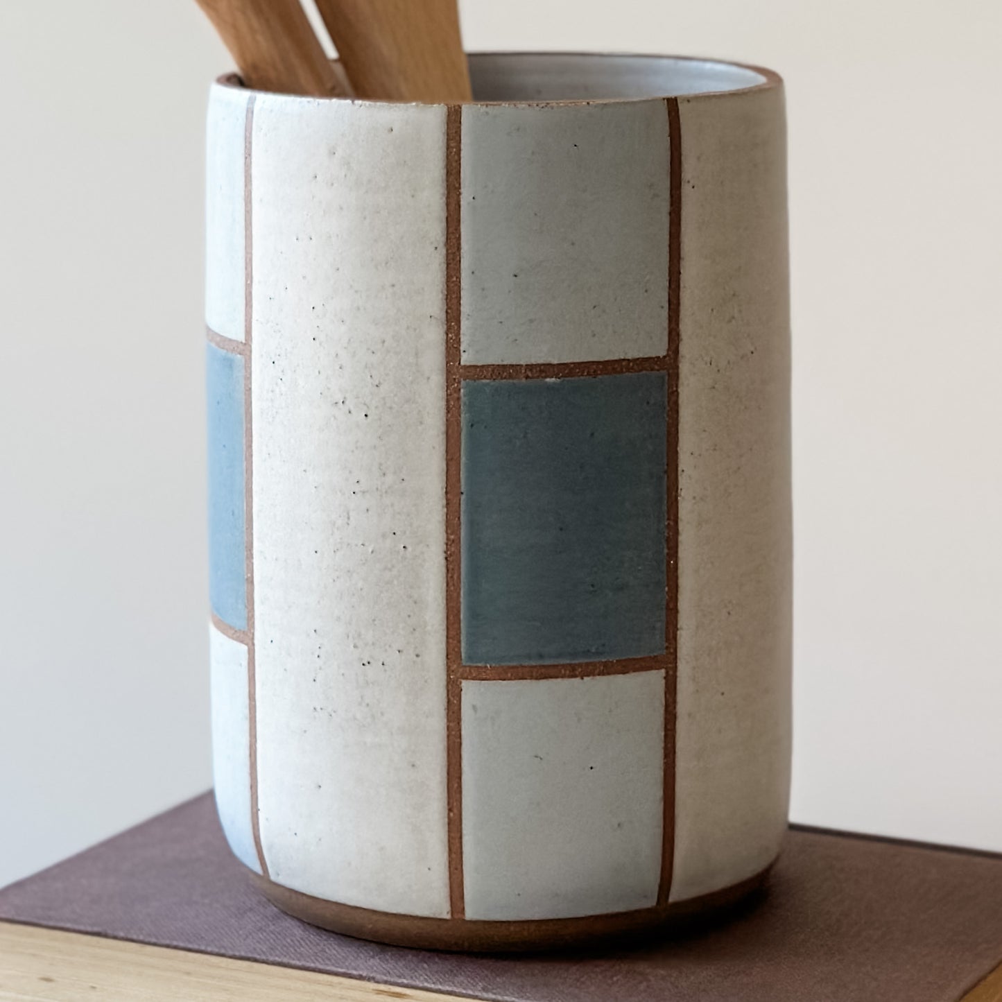 A close up view of a geometric handmade ceramic utensil holder sitting on a book. The utensil holder is dark blue, grey and white.