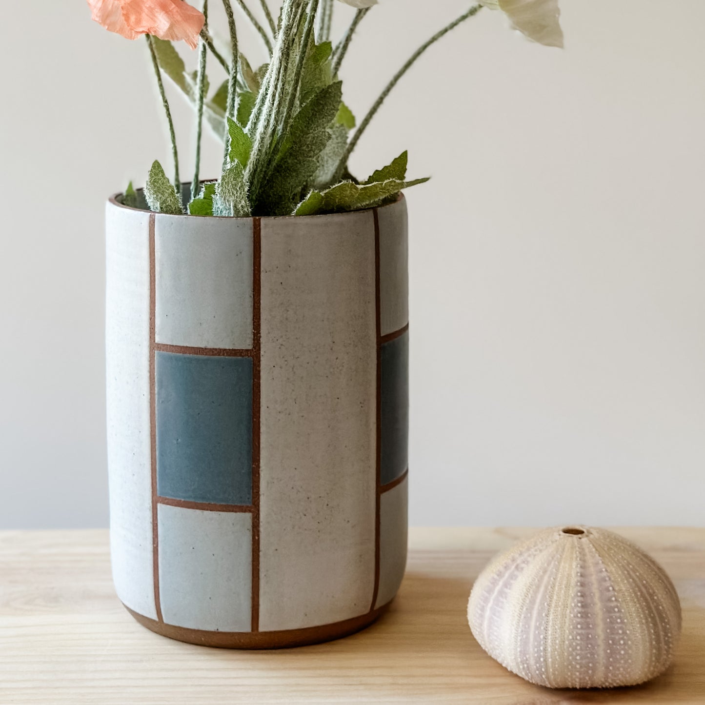 A close up view of the geometric handmade ceramic vase holding flowers. The vase sits on a wooden table with a seashell. The vase is dark blue, grey and white.
