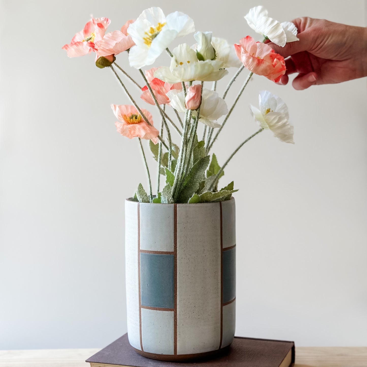 A geometric handmade ceramic vase with flowers on a book on a wooden table. The vase is dark blue, grey and white. A hand is arranging flowers in the vase.