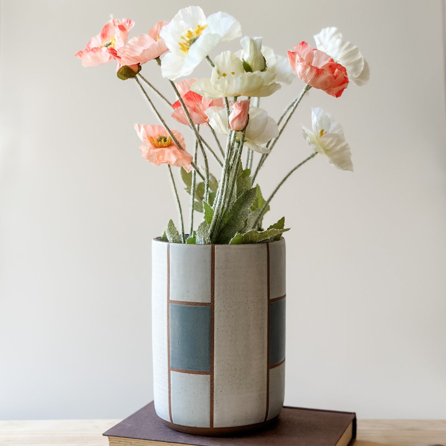 A geometric handmade ceramic vase sits on a book on a wooden table. The vase is dark blue, grey and white. The vase holds flowers in pastel colours.