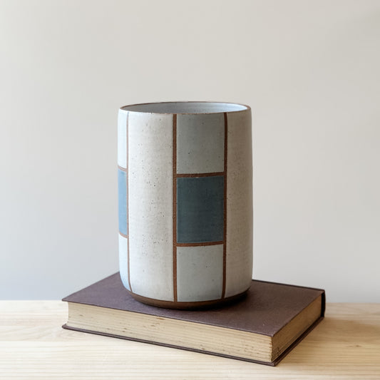 A geometric handmade ceramic utensil holder sits on a book on a wooden table. The utensil holder is dark blue, grey and white.