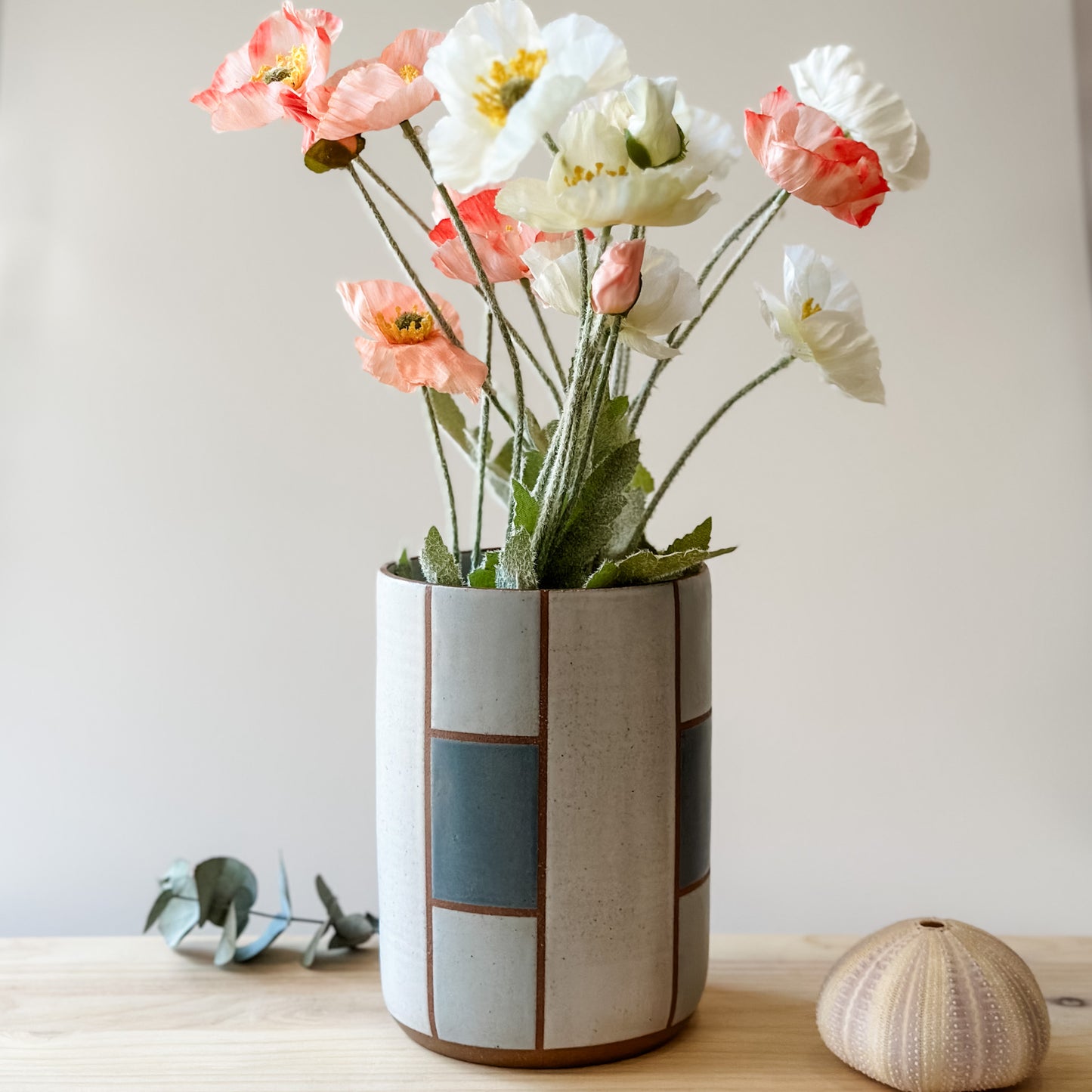 A geometric handmade ceramic vase sitting on a book on top of a wooden table. The vase is hand-painted in dark blue, grey and white. The handmade vase holds flowers.