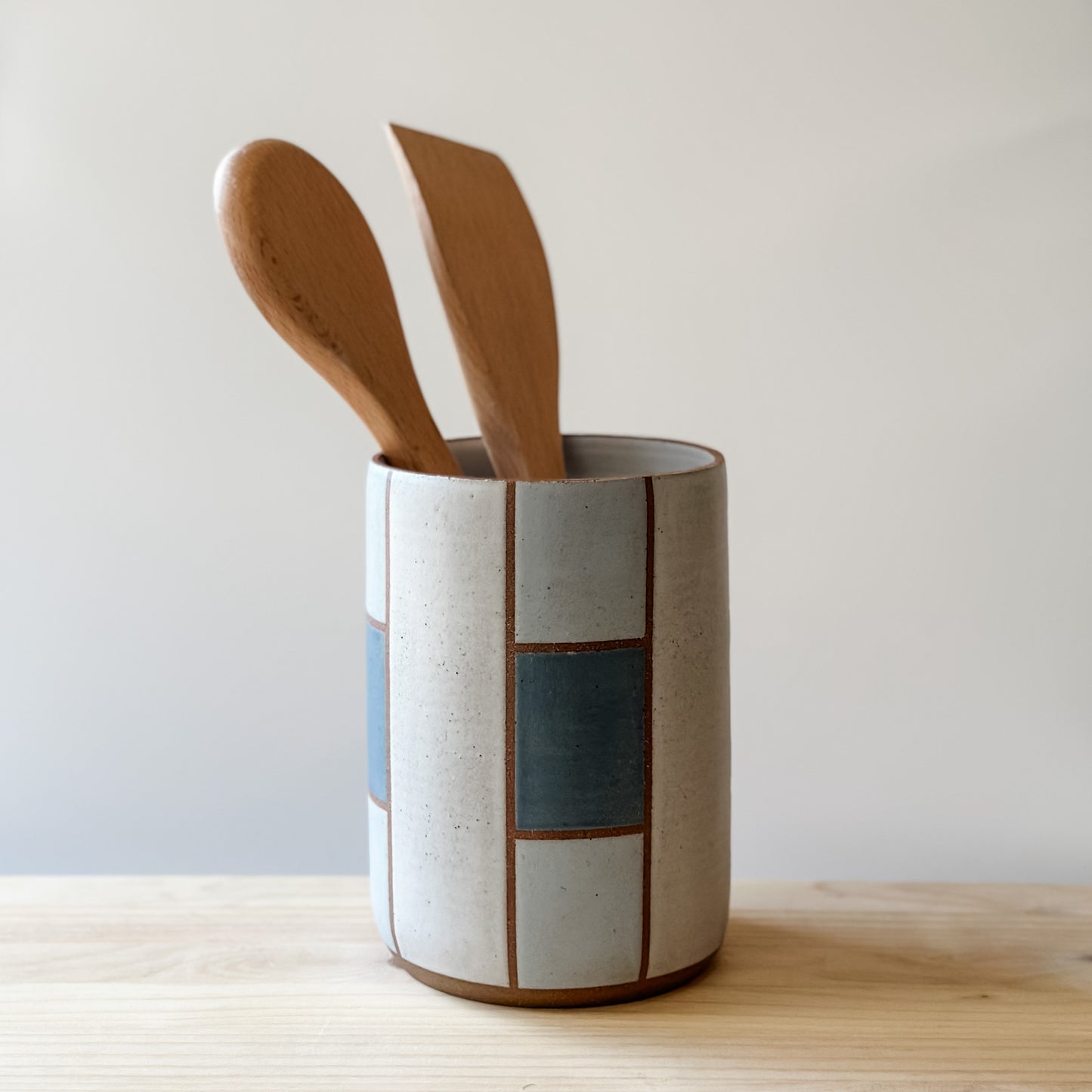 A geometric handmade ceramic utensil holder sits on a book on a wooden table. The utensil holder is hand-painted in dark blue, grey and white. The handmade utensil holder holds a wooden spoon and spatula.