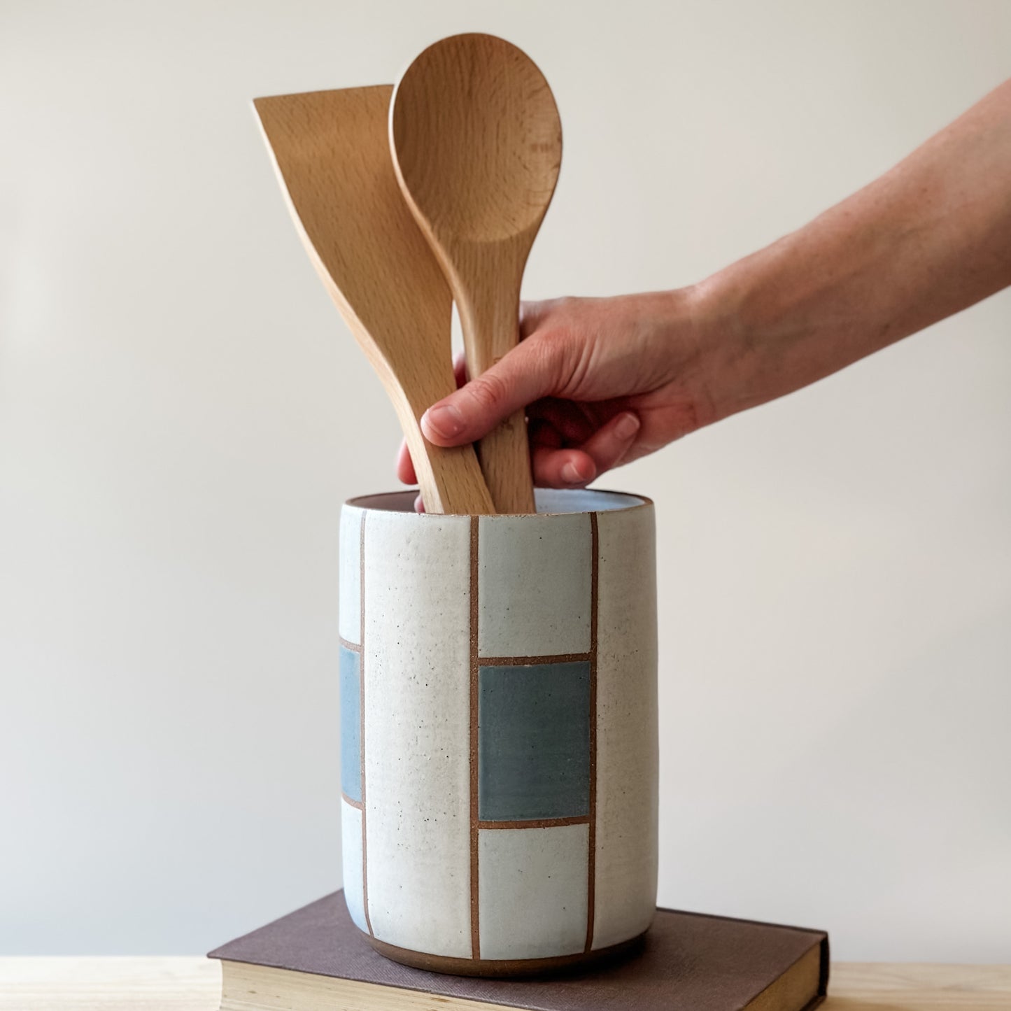 A geometric handmade ceramic utensil holder sits on a book on a wooden table. The utensil holder is dark blue, grey and white. A hand is placing wooden utensils into the utensil holder.