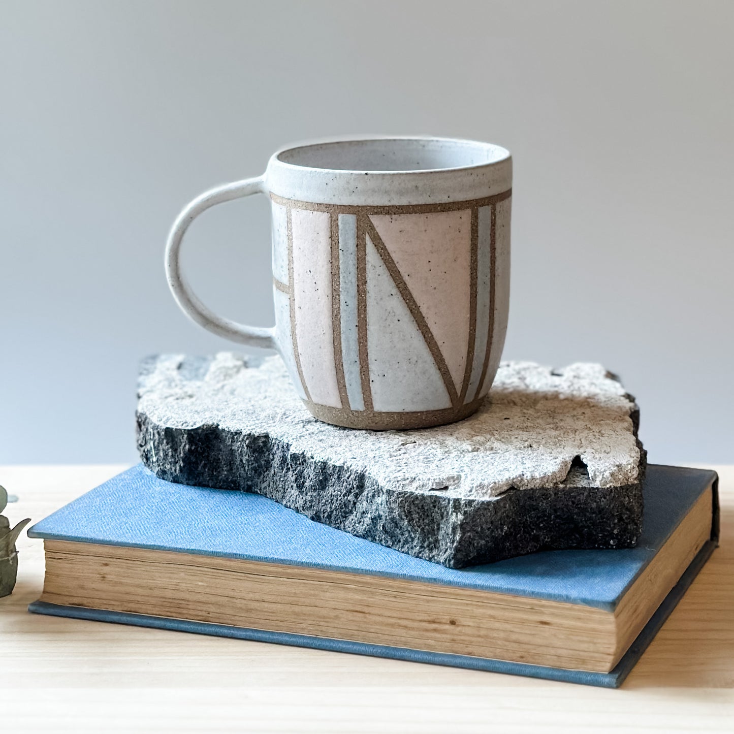 A front view of the limited edition Birdrock Ceramics geometric mug. The mug is painted pink, white and grey. The mug sits on a tile, a book, and a wooden table.