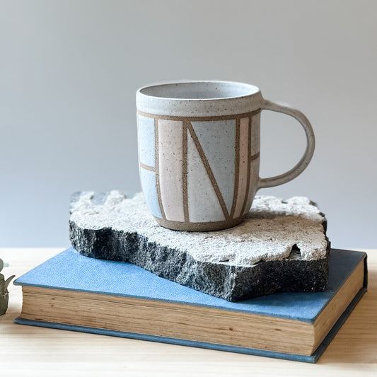A front view of the limited edition Birdrock Ceramics geometric mug. The mug is painted pink, white and grey. The mug sits on a tile, a book, and a wooden table.