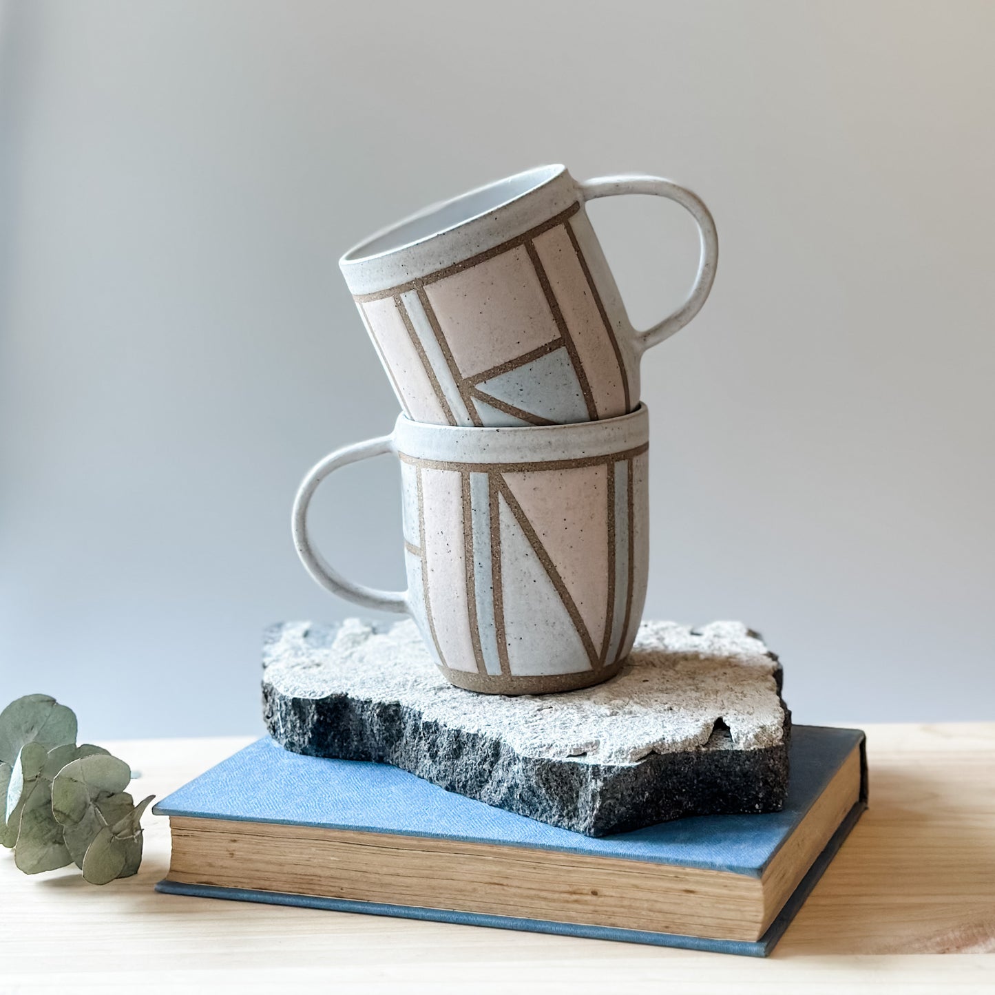 A front view of two limited edition Birdrock Ceramics geometric mugs. The mugs are painted pink, white and grey. The mugs sit on a tile, a book and a wooden table.