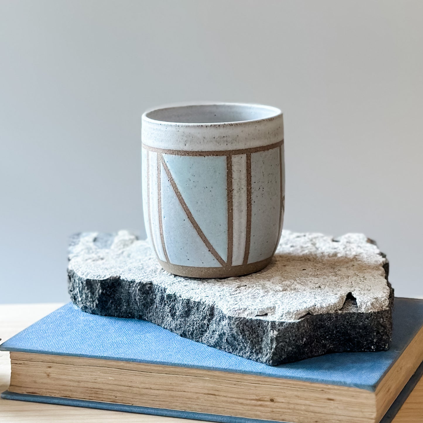 One limited edition geometric handmade ceramic tumbler sits on a tile and a book. It is glazed in light blue, white and grey glaze.