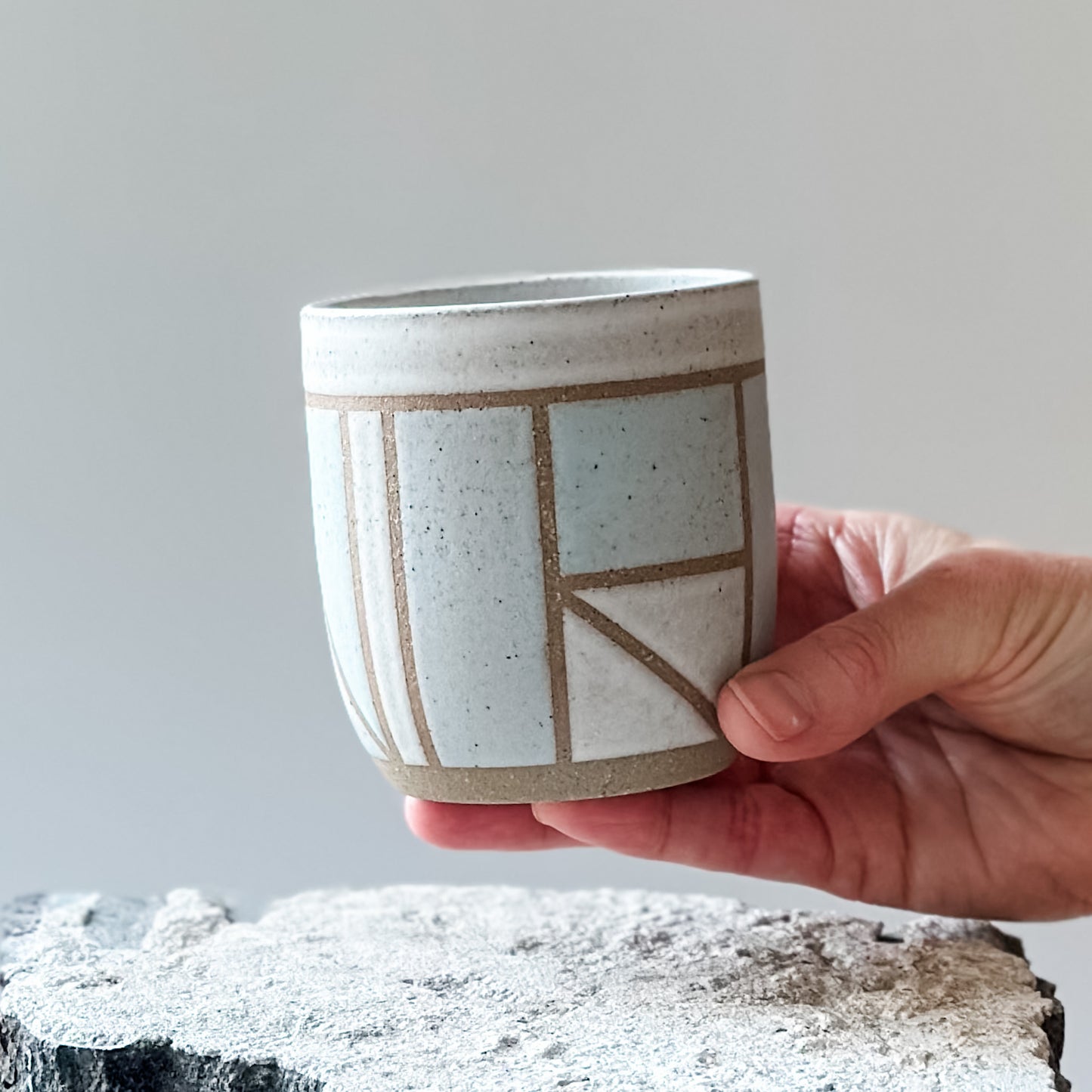 A limited-edition geometric handmade ceramic tumbler. It is held above a tile and glazed in light blue, white and grey glaze.