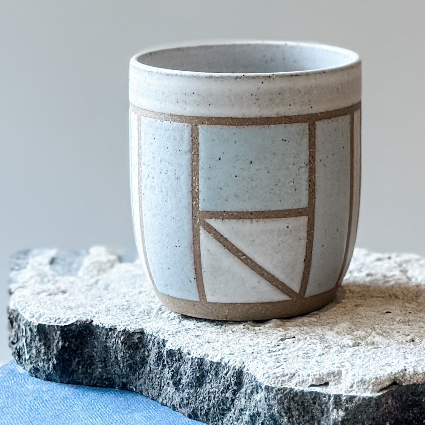 One limited edition geometric handmade ceramic tumbler sits on a tile. It is glazed in light blue, white and grey glaze.