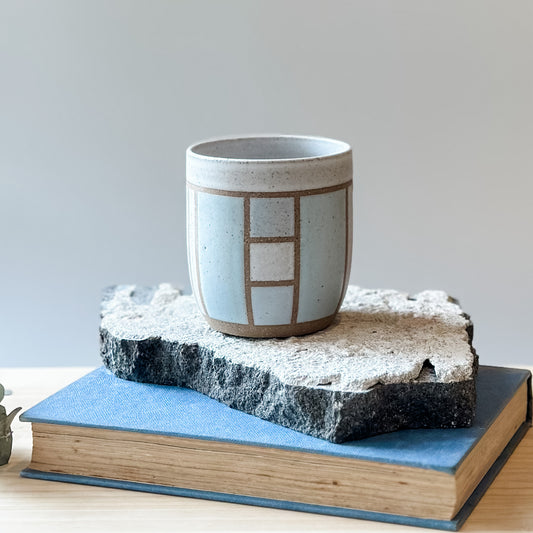 One limited edition geometric handmade ceramic tumbler sits on a tile and a book. It is glazed in light blue, white and grey glaze.