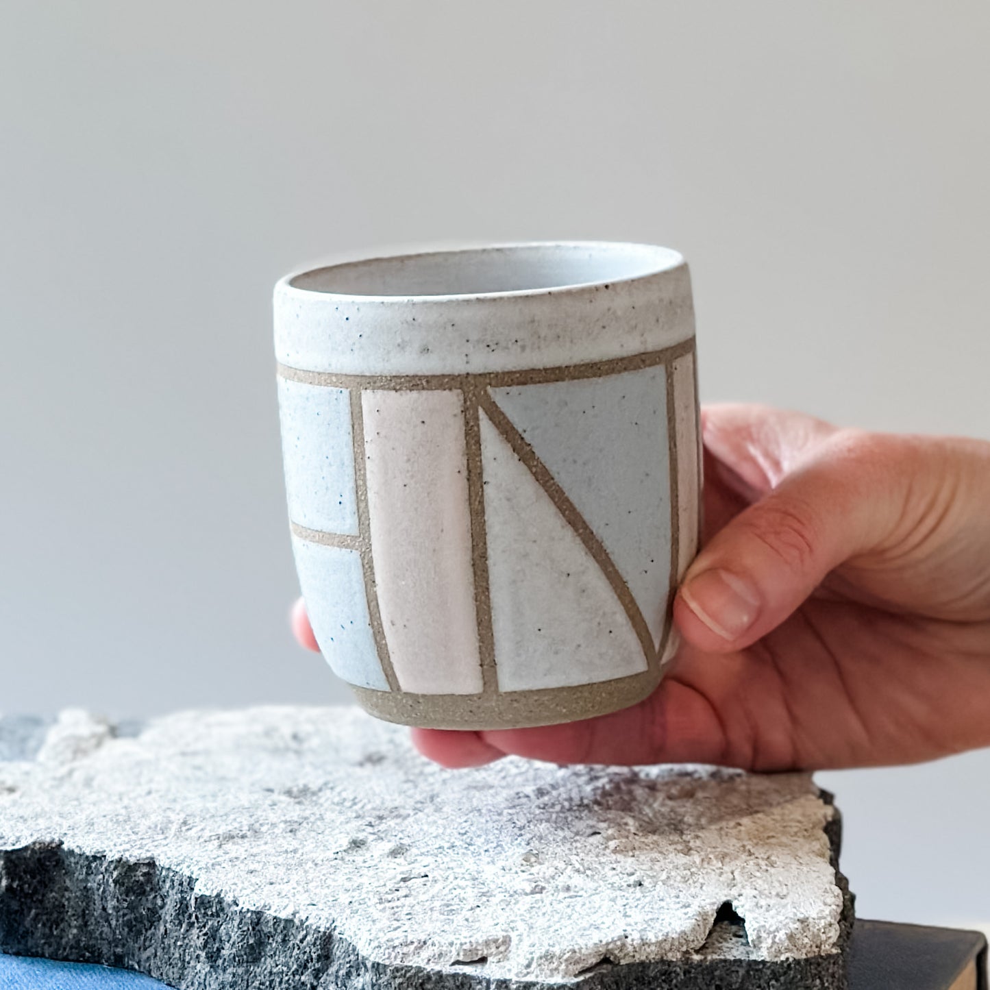 One handmade ceramic tumbler with a geometric pattern. It is held above a tile and is glazed pink, grey and white.