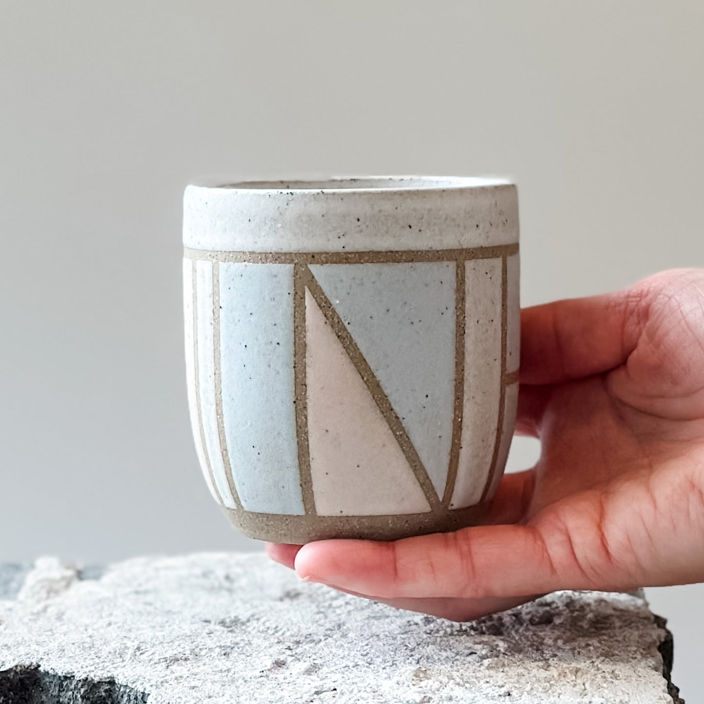 One handmade ceramic tumbler with a geometric pattern. It is held above a tile and is glazed pink, grey and white.