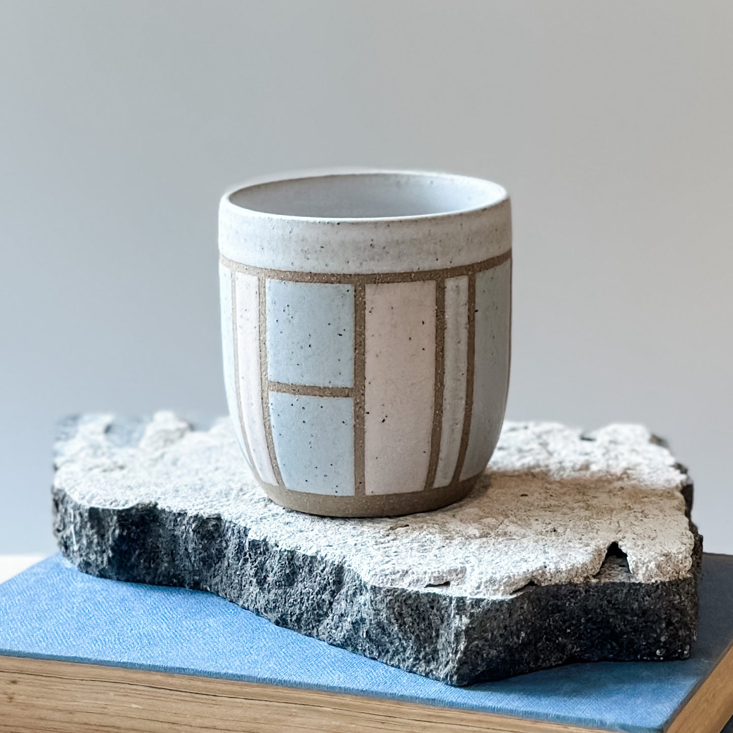 One handmade ceramic tumbler with a geometric pattern sits on a tile and a book. It is glazed pink, grey and white.