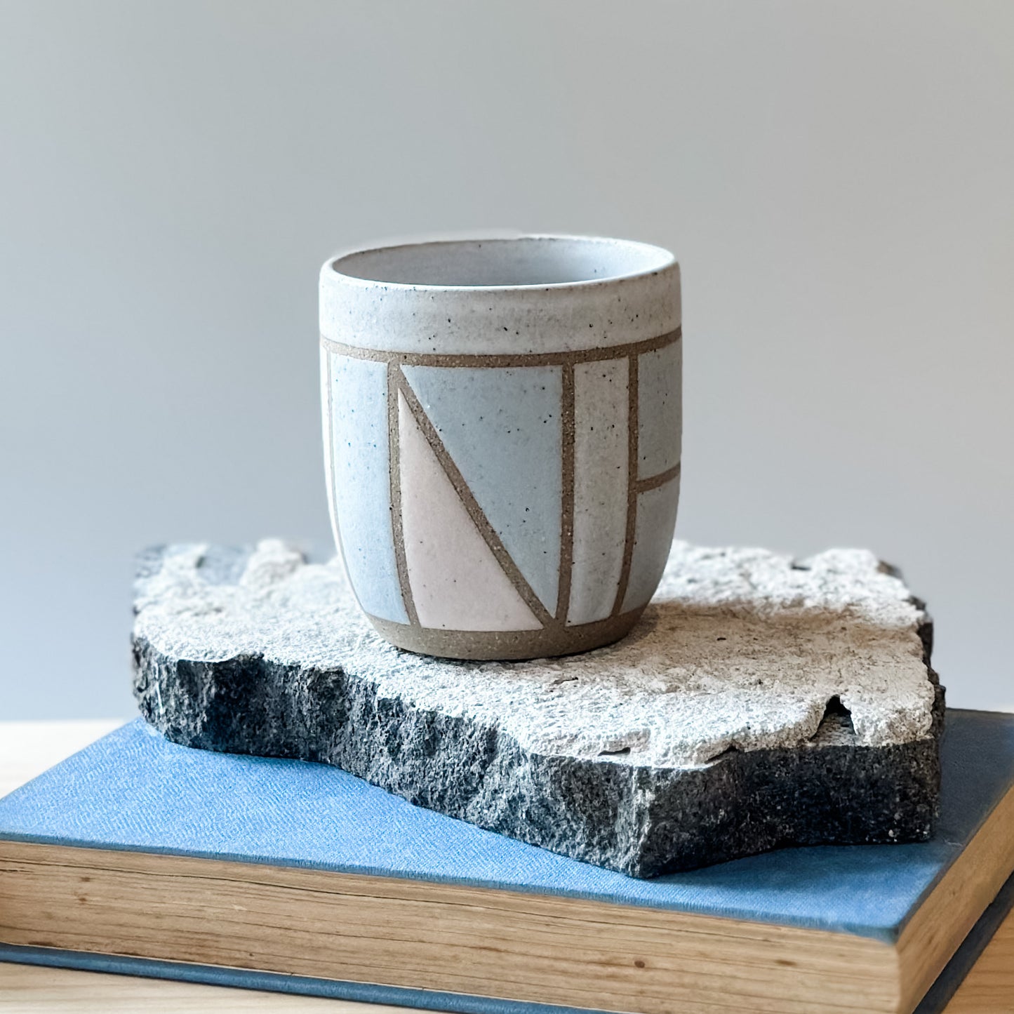 One handmade ceramic tumbler with a geometric pattern sits on a tile and a book. It is glazed pink, grey and white.