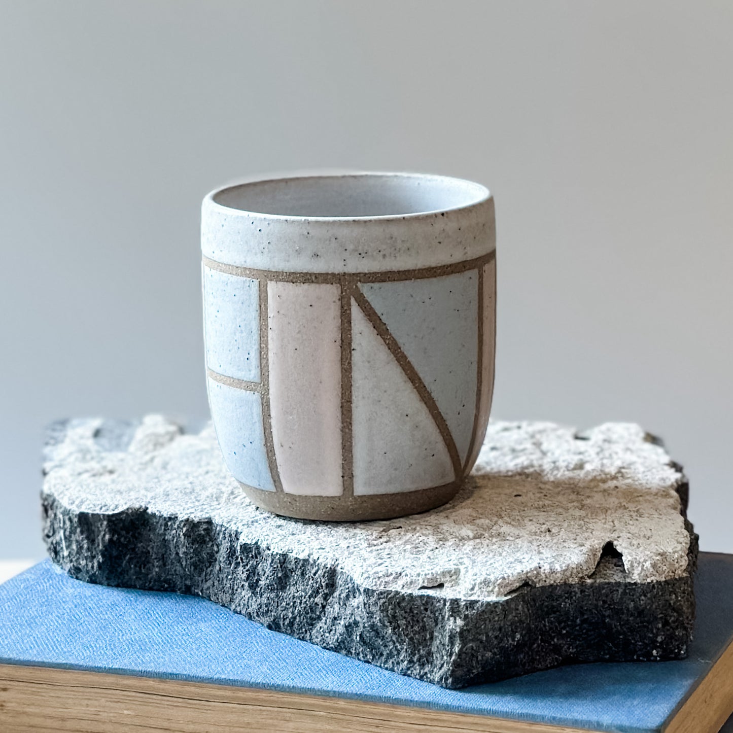 One handmade ceramic tumbler with a geometric pattern sits on a tile and a book. It is glazed pink, grey and white.