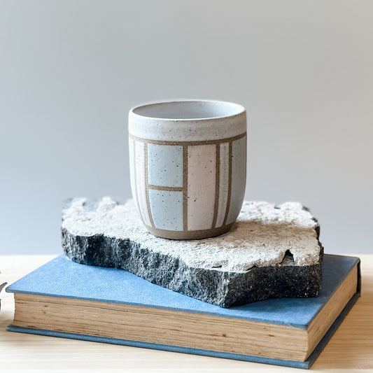 One handmade ceramic tumbler with a geometric pattern sits on a tile and a wooden table. It is glazed pink, grey and white.