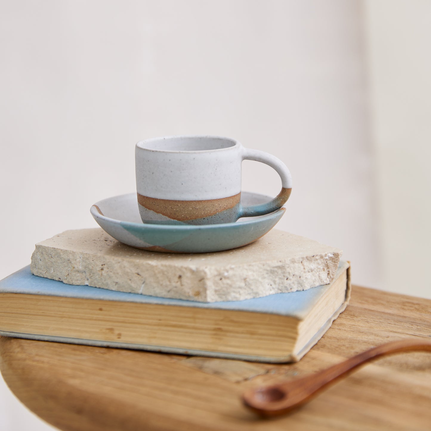 A front view of the 'Tidal Handmade Ceramic Espresso Mug'. The handmade espresso mug has teal green, white and grey glaze. The espresso mug is on a tile, book and wooden table in a coastal setting. The Espresso Mug is pictured with the Espresso Saucer.