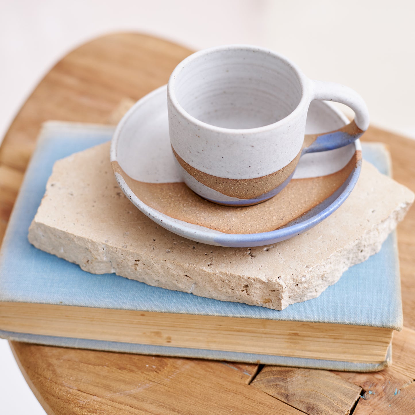 A top view of the 'Tidal Handmade Ceramic Espresso Mug'. The handmade espresso mug has violet, white and grey glaze. The espresso mug is pictured with an espresso saucer on a tile, book and wooden table in a coastal setting.