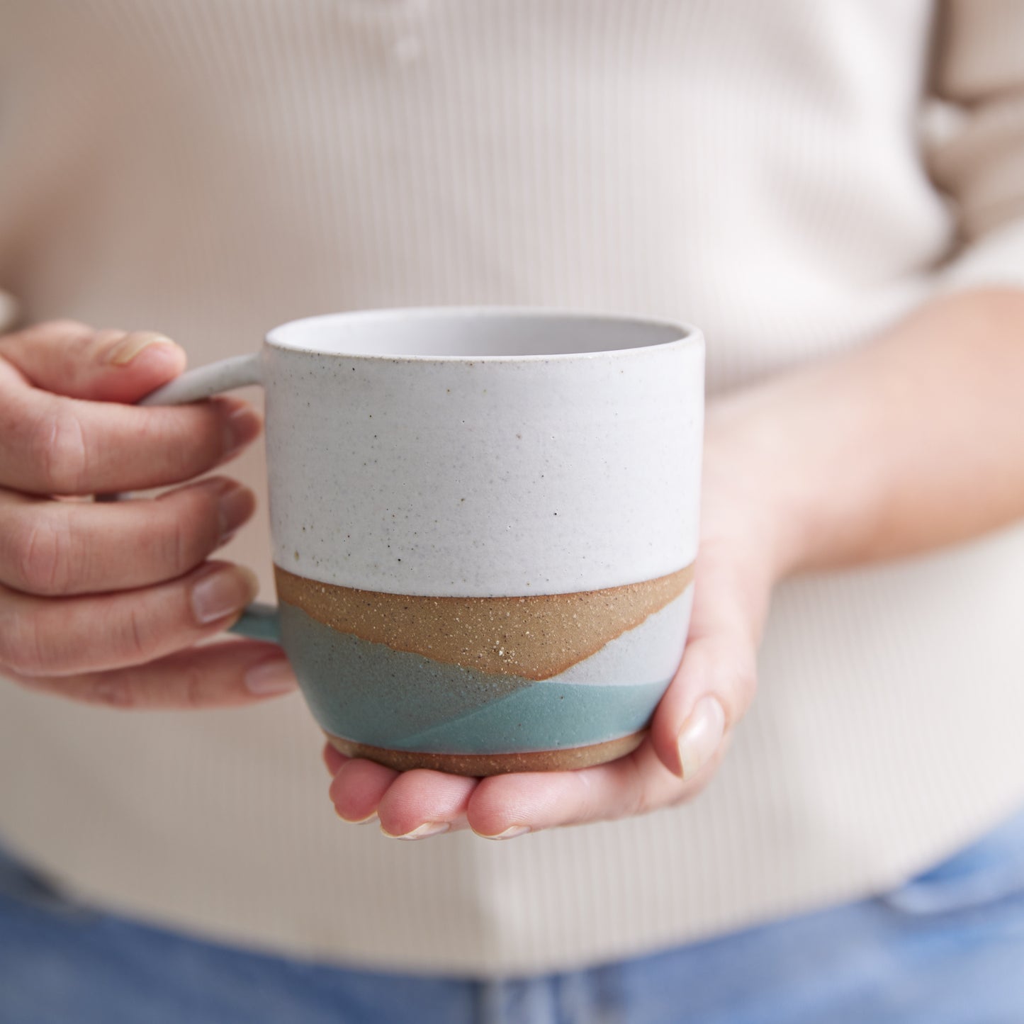 A handmade ceramic mug being held in two hands. The mug has a white, green and grey glaze.