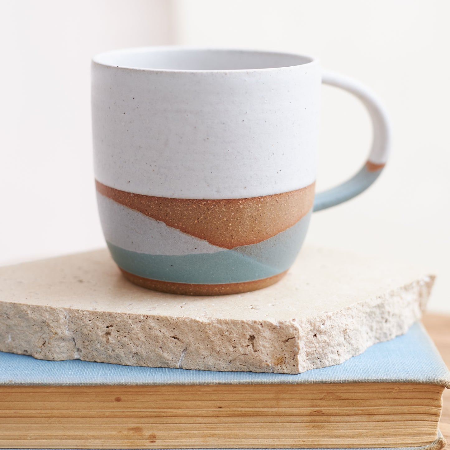 A close up image of a handmade ceramic mug in a white, green and grey glaze. The handmade mug is sitting on a tile and book in a coastal setting.