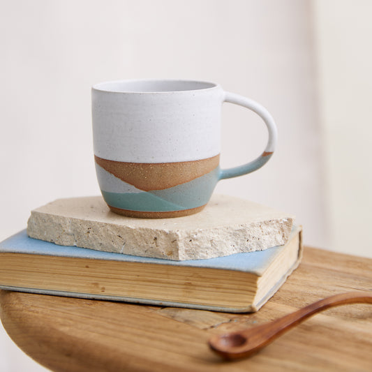 A handmade ceramic mug in a white, green and grey glaze. The ceramic mug is sitting on a book and wooden side table in a coastal setting.