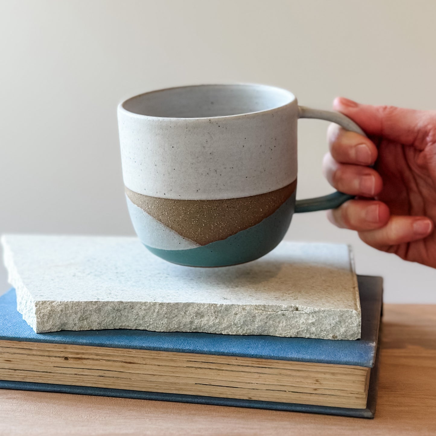 Hand holding a ceramic mug with a coastal design on top of two stacked books.