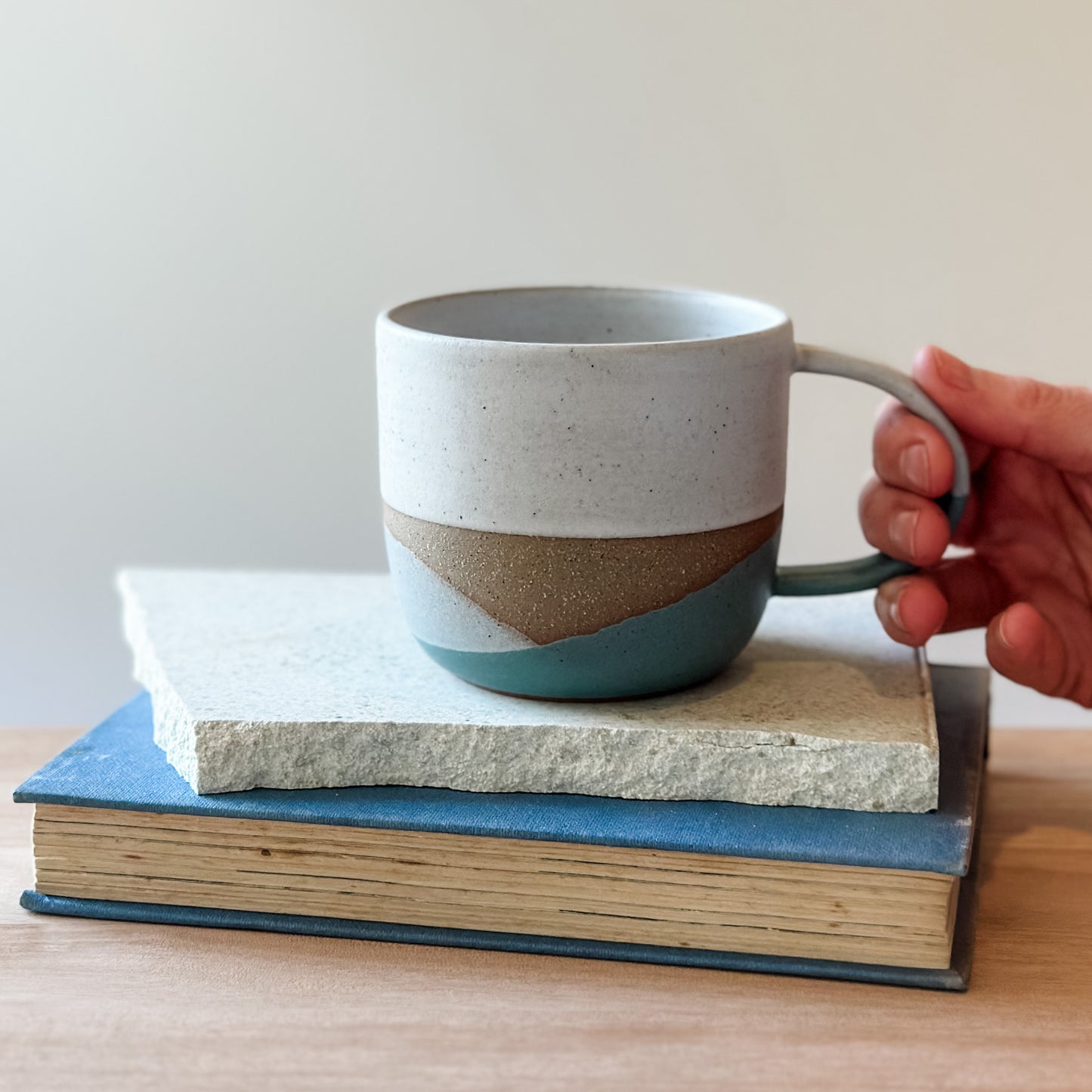 Hand holding a ceramic mug with a coastal design on top of a tile and a book.