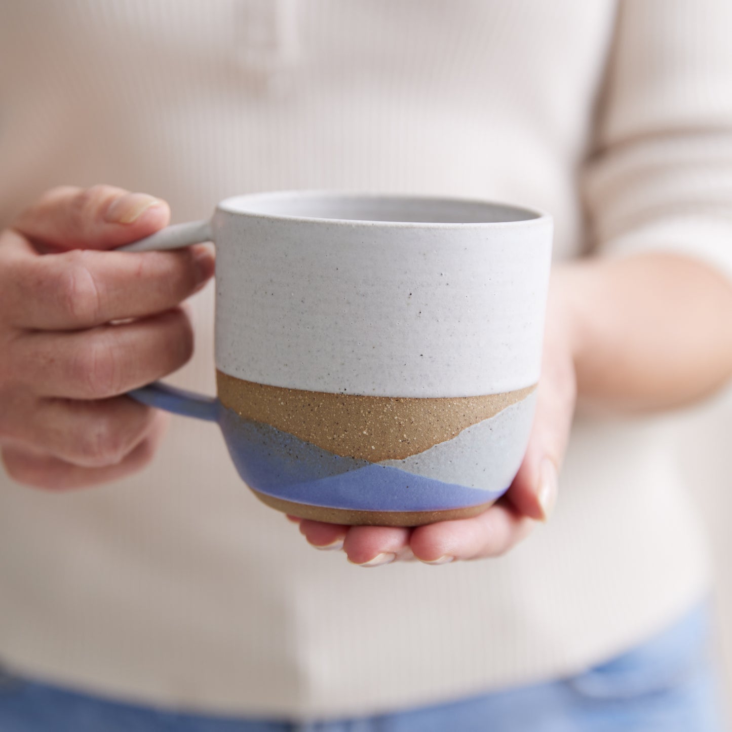 A Handmade Ceramic Mug being held in two hands. The handmade mug has a purple, grey, and white glaze.