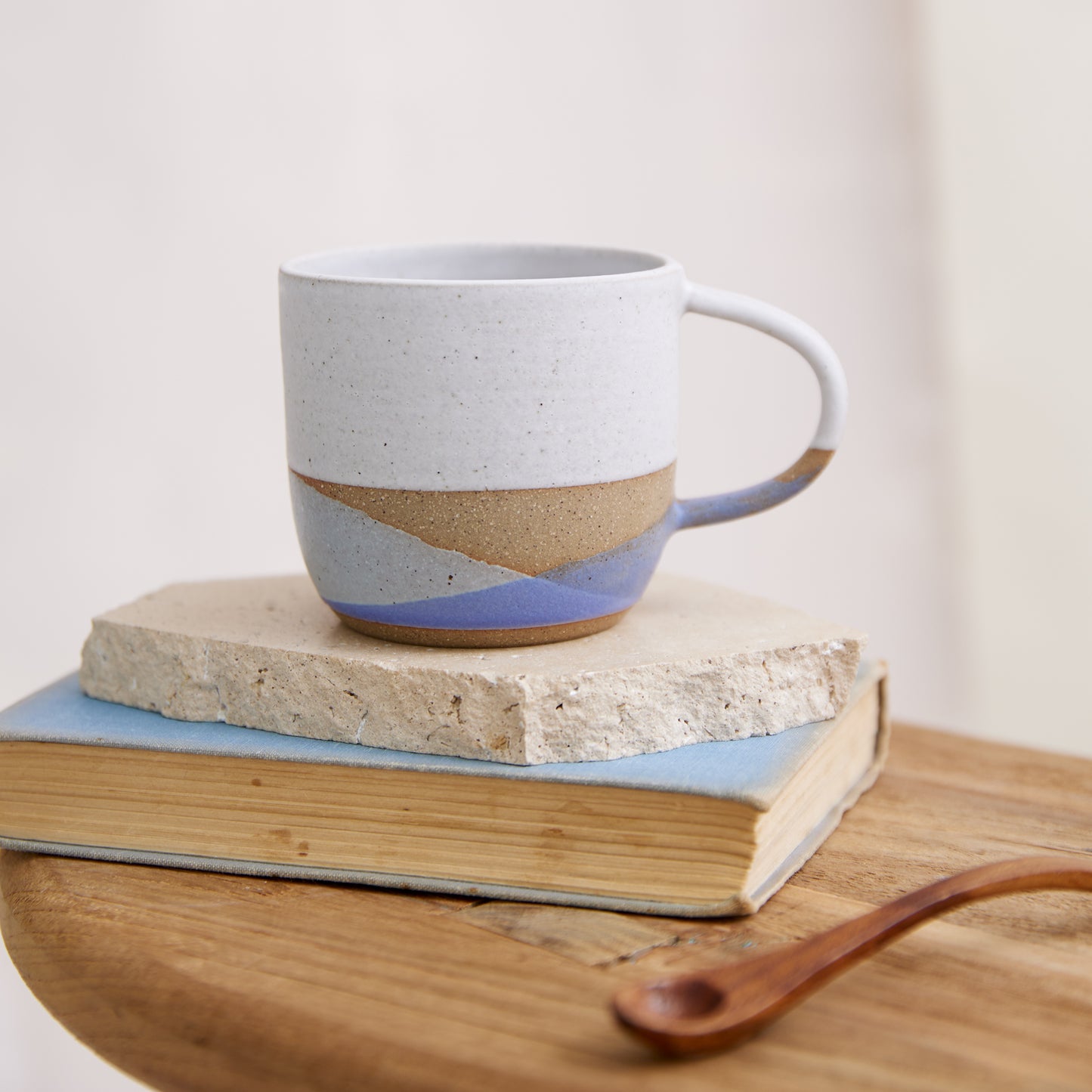 A handmade ceramic mug in a white, purple and grey glaze. The handmade mug is sitting on a tile, on top of a book and wooden table in a coastal setting.