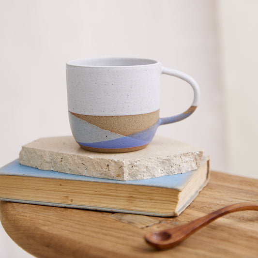 A handmade ceramic mug in a white, purple and grey glaze. The handmade mug is sitting on a tile, on top of a book and wooden table in a coastal setting.