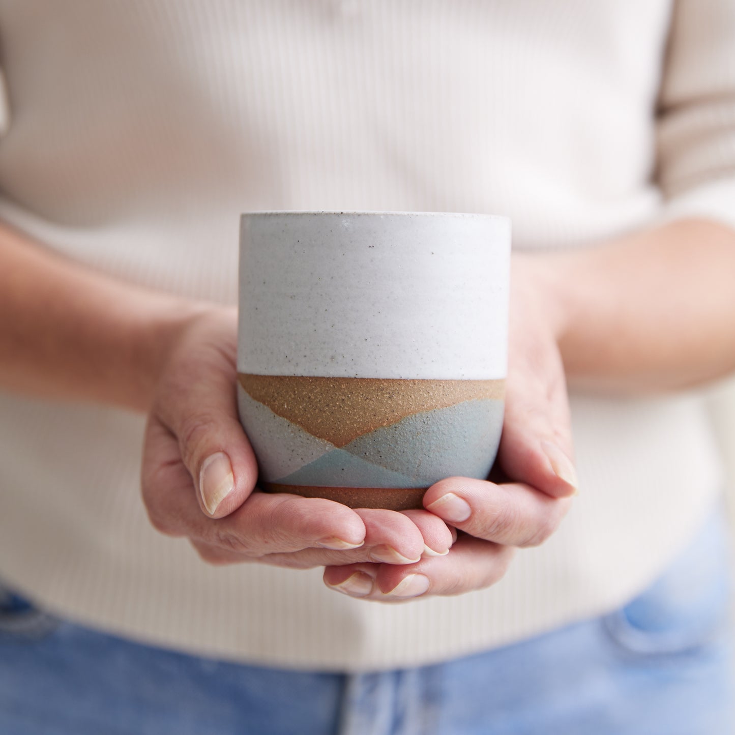 A Handmade Ceramic Tumbler in a grey, green, and white glaze. The ceramic cup is being held in two hands.