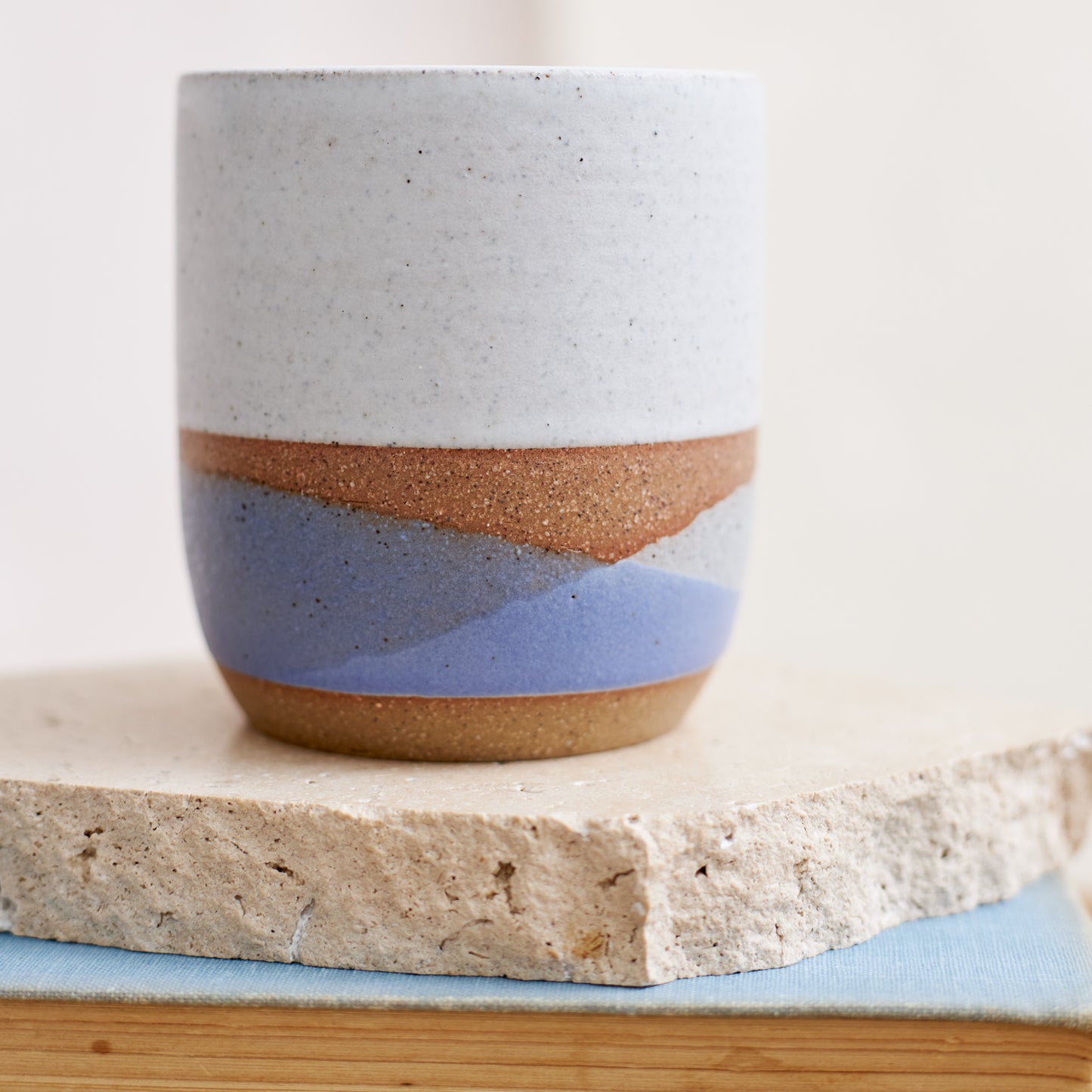 A close up image of a handmade ceramic tumbler sitting on a book and tile in a coastal setting. The handmade ceramic tumbler is glazed in a white, violet and grey glaze.