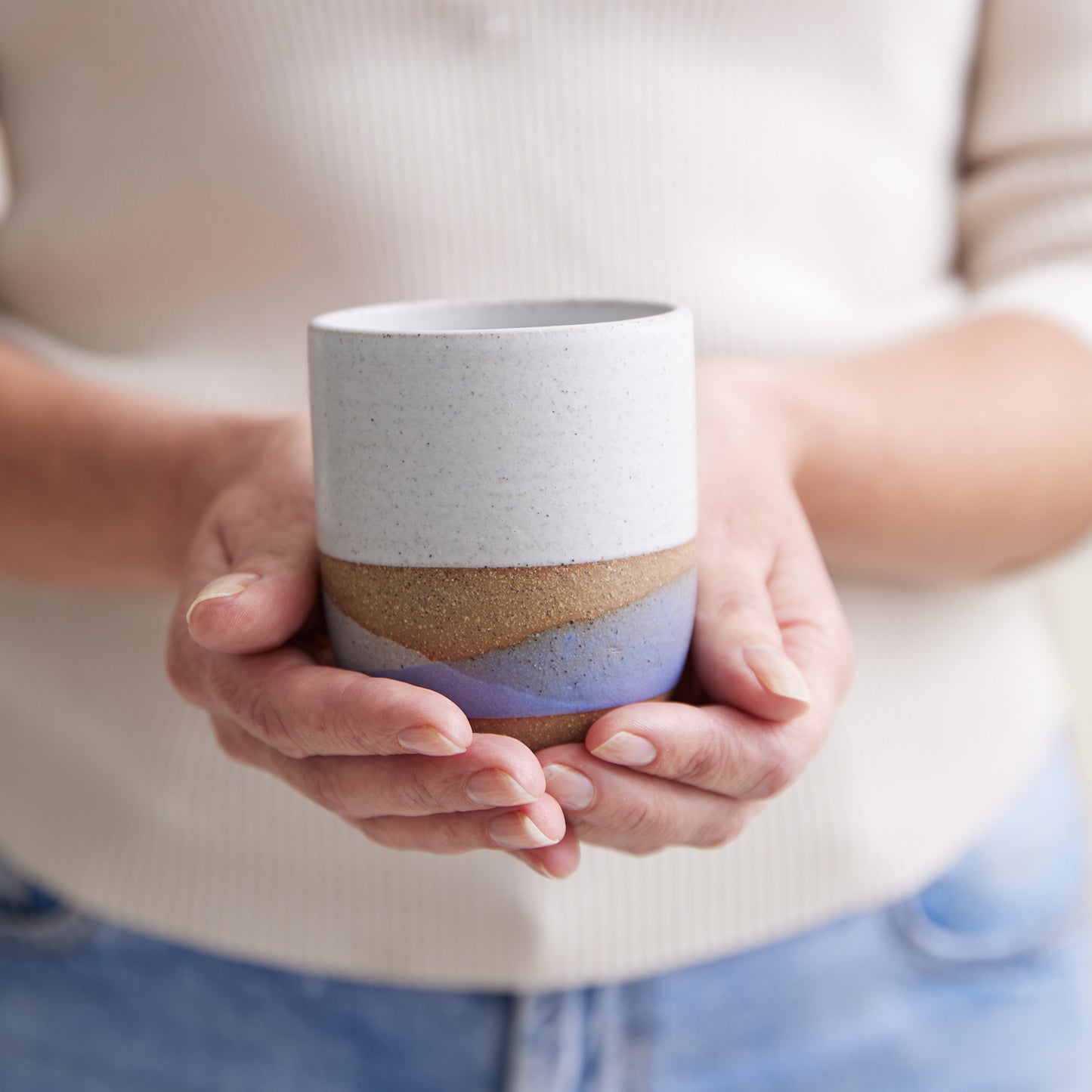 A handmade ceramic tumbler being held by two hands. The handmade ceramic tumbler is glazed in a white, violet and grey glaze.