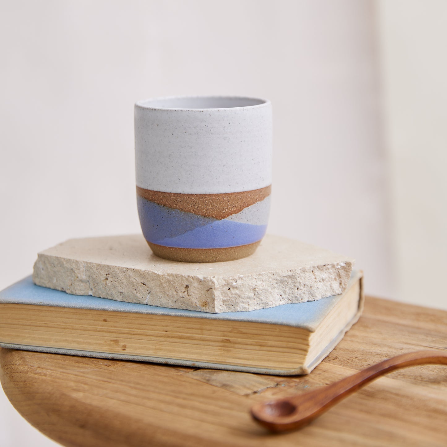 A handmade ceramic tumbler sitting on a book and side table. The handmade ceramic tumbler is glazed in a white, violet and grey glaze.